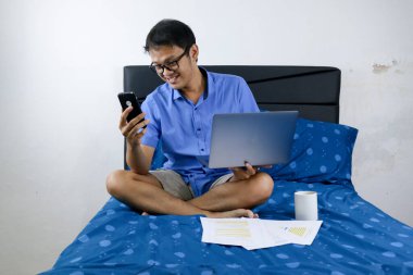 Genç Asyalı adam evde dizüstü bilgisayar ve telefonla çalışırken gülümsüyor ve eğleniyor. Yatakta oturup çalışan bir adam. Ev kavramından çalış.