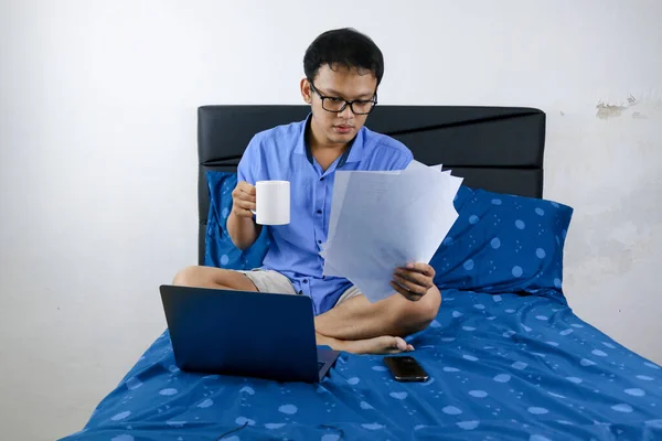 Young asian man is working at home wearing glasses. A man sitting on bed and working with laptop computer and document with drinking a cup of coffee on bed. Work from home concept.