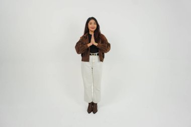 Asyalı genç bir kadının vücut portresi. Sıradan kıyafetleriyle iki eliyle selam veriyor. Gülümsüyor, izole beyaz arka planda duruyor.