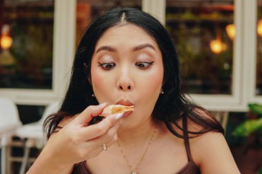 Joyful young Asian woman in brown dress eating a slice of pizza at a restaurant.