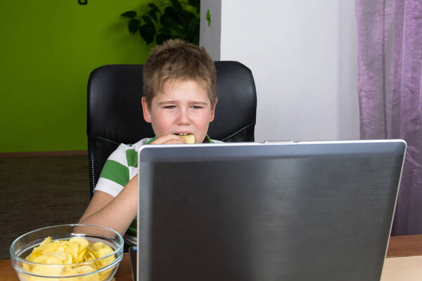 Obese little boy sitting at the computer and eating chips.