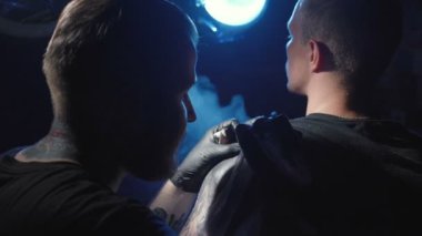 Genç bir adamın omzuna dövme yaptıran bir profesyonelin görüntüsü.