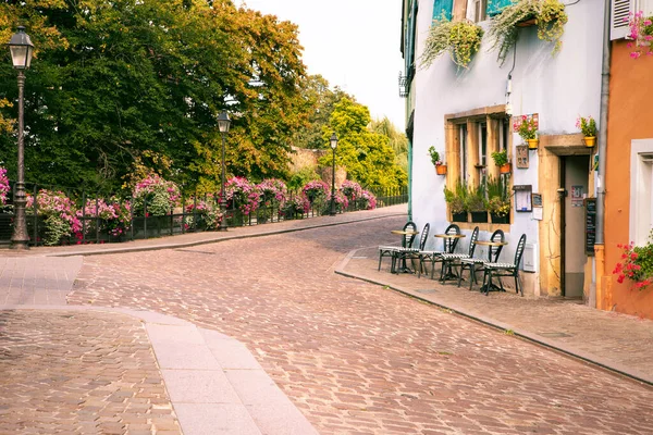 View on romantic french cobblestone street with cafe, street lamps and park in background. Empty tables and chairs outside the restaurant by the facade of colourful buildings in Colmar, Alsace, France