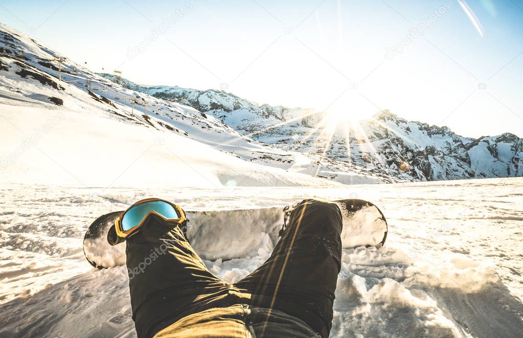 Snowboarder sitting at sunset on relax moment in european alps ski resort - Winter sport concept with guy and snowboard on mountain top ready to ride - Legs point of view on vintage contrast filter