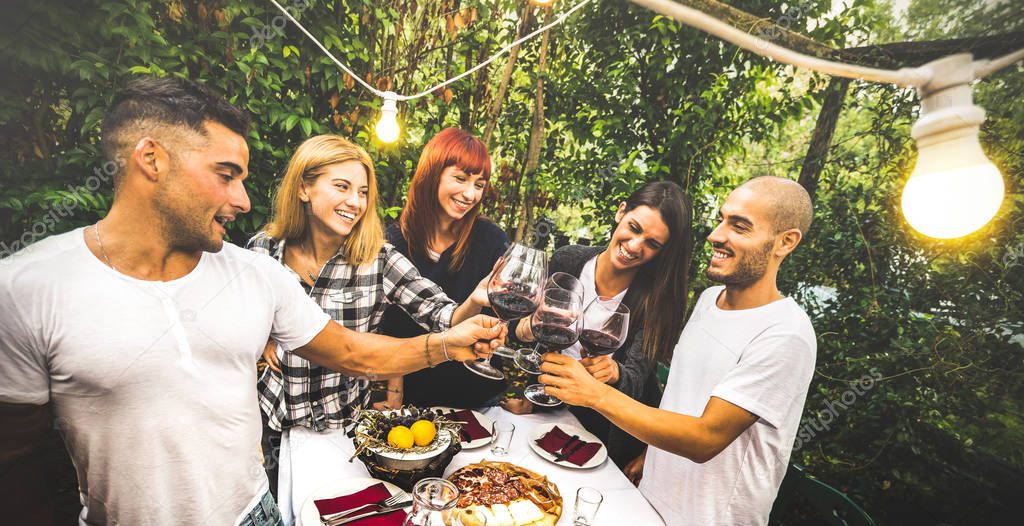 Happy friends having fun drinking red wine at backyard garden party - Youth friendship concept together at farm house vineyard winery - Focus on background young women with bulb lights lighting on men