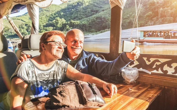 Casal aposentado sênior de turistas felizes tirando selfie na excursão de exploração Mekong com barco lento no Laos PDR - Conceito de viagem idoso ativo em viagem ao redor do mundo - Filtro de pôr do sol saturado quente — Fotografia de Stock