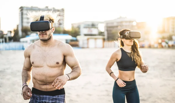 Pasangan muda yang berlari di pantai dengan kacamata virtual reality headset Olahraga dan teknologi konsep dengan teman-teman joging dan berolahraga di perjalanan simulasi pada musim panas Kontras matahari terbenam filter — Stok Foto