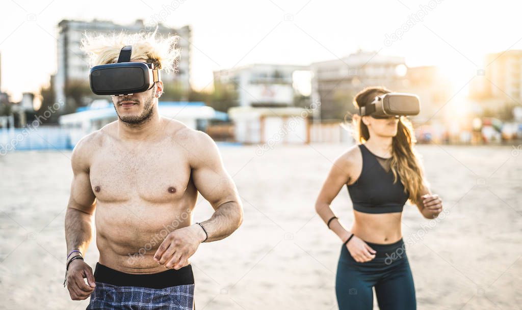 Young couple running at beach with virtual reality goggles headset - Sport and technology concept with friends jogging and exercising on trek simulation on summer time - Contrast sunset filter