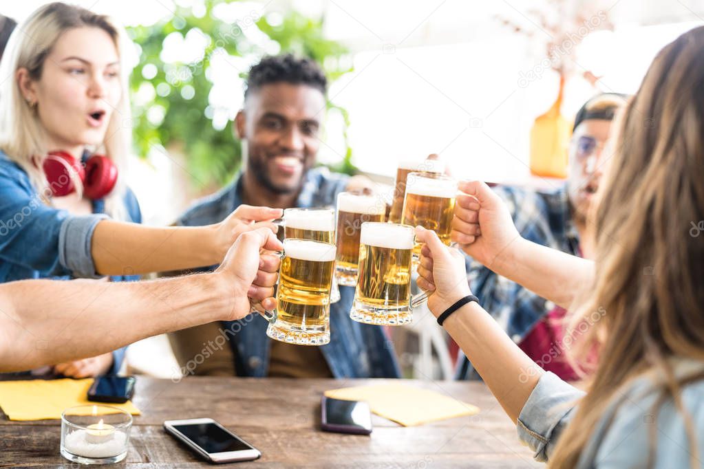 Happy multiracial friends drinking and toasting beer at brewery bar - Friendship concept with young people having fun together at cool pub restaurant - Focus on middle pint glasses - Bright filter