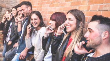 Milenyum erkekleri ve kızları cep telefonu kullanıyor ve şehirdeki tuğladan taş duvarda eğleniyorlar - genç milenyum insanlarıyla şehir gençliği ve teknoloji konsepti sıcak vintage filtrede arkadaşlar