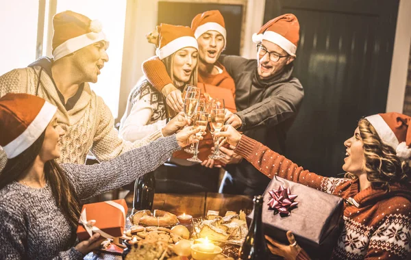 Friends group with santa hats exchanging Christmas presents with champagne wine toast at home dinner - Winter holiday concept with young people enjoying time and having fun together - Focus on glasses — Stock Photo, Image