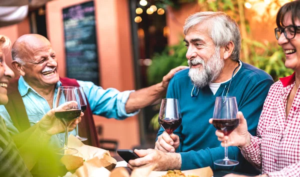 Happy senior friends having fun drinking red wine at dinner party - Retired people eating at restaurant together - Dinning lifestyle concept on warm vivid filter - Focus on bearded hipster man