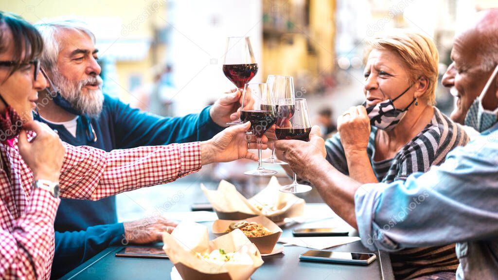 Senior couples toasting red wine at restaurant bar with face masks - New normal lifestyle concept with happy people having fun together at bar outdoors - Bright filter with focus on central glasses
