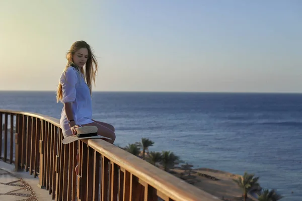一个性感的美丽的晒黑的女人在泳装和浅海滩裙在阳台上与蓝色大海和天空的背景的肖像。暑假概念. — 图库照片