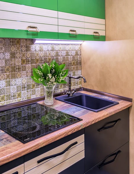 Modern kitchen in retro style. A bouquet of lilies of the valley in a vase stands on a wooden tabletop next to an induction cooker and sink