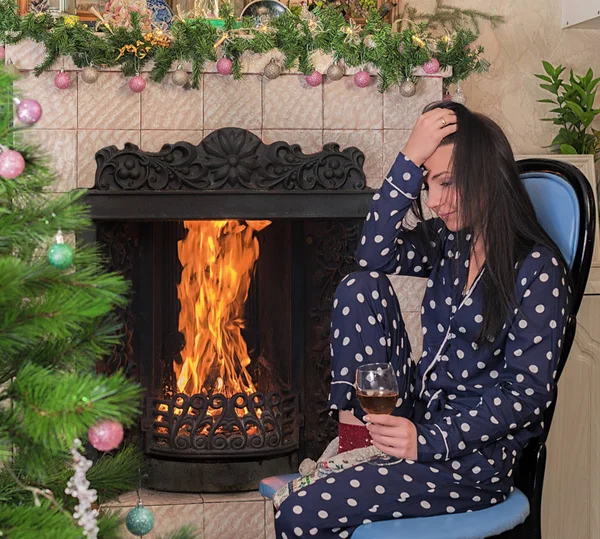 A young brunette girl is sitting by the fireplace in the living room and holding a glass of wine in her hand. New Year, Christmas, Christmas tree and festively decorated interior.