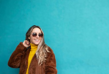 Uzun sarı saçlı gülümseyen neşeli kız. Sarı kazaklı, sahte kürklü ve güneş gözlüklü Avrupalı kız. Sonbahar giysileri. Mavi arkaplanda genç bir kadın ve metin için boşluk