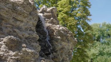 Parkta şelale. Akan su. Taşlardan su dökülür ve düşer. Mavi gökyüzü ve yeşil ağaçların arka planında. Güneşli bir gün. Çeşme.