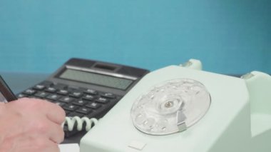 Telefon konuşmaları. Eski bir cihaz. Alıcı kapalı. El bir kalem tutar, hesap makinesine güvenir ve veri yazar.