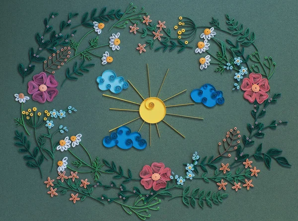 The sun with clouds inside round frame with paper flowers and leaves isolated on a green paper background. Top view. Hand made of paper quilling technique.