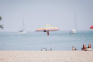 Sahilde güneşli bir günün bulanık arka planı, kıyı şeridini süsleyen renkli şemsiyeler insanlar güneşin sıcaklığının ve denizin yumuşak dalgalarının tadını çıkarırken