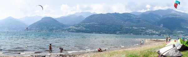 Colico, İtalya - 21 Temmuz 2019: Como gölündeki plajın panoramik manzarası. Kitesurfer bir hile yapma, çocuklar bir plajda oynarken, bir zemin üzerinde kurutma ekipmanları kiting. Arka planda Alp dağları. .