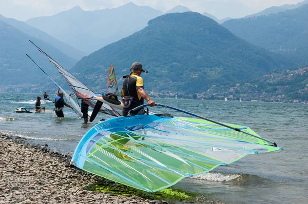 Lago de Como, Italia - 21 de julio de 2019. Hombre windsurfer llevando una vela en el lago de la montaña, tabla de surf en primer plano. Montañas de los Alpes sobre un fondo — Foto de Stock