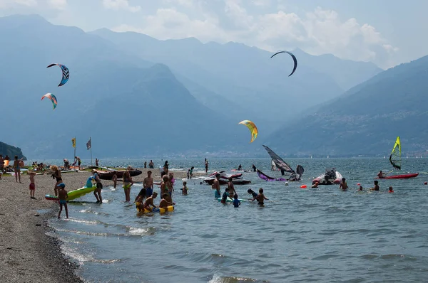 Lago de Como, Italia - 21 de julio de 2019: Personas nadando en el lago de montaña. Niños jugando. Entrenamiento de kitesurfistas y windsurfistas. Árboles y montañas de los Alpes en el fondo — Foto de Stock