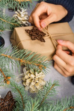 Kadın eli, gri arka planda bir hediye kutusu tutuyor. Kozalaklı ve ladin dallı kompozisyon. Bayramlar için şenlikli arka plan: Noel, Yeni Yıl. Düz yatıyordu.