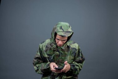 The young man wears a camouflage raincoat and shows different gestures. On a gray background in the studio.
