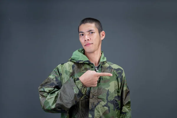 The young man wears a camouflage raincoat and shows different gestures. On a gray background in the studio.