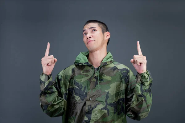 The young man wears a camouflage raincoat and shows different gestures. On a gray background in the studio.