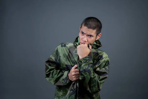 The young man wears a camouflage raincoat and shows different gestures. On a gray background in the studio.