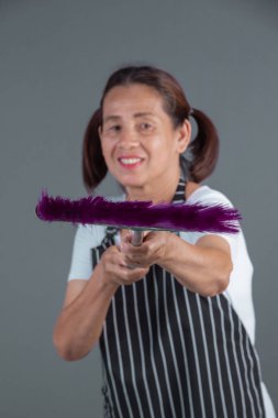 Yaşlı bir ev hanımı, elinde gri bir arka planda plastik süpürge tutuyor..