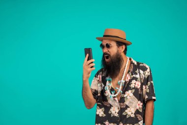 Uzun ve mutlu bir sakallı adamın şapka giymesi, çizgili gömlek giymesi, mavi arka planda telefon tutması..