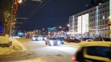 Gece şehri, kış şehri. Tatil için dekore edilmiş arabalar belediye binasının önünden geçiyor. Kış. Novosibirsk.