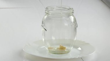 Vejetaryen, basit yemek. Beyaz bir masada duran şeffaf cam bir kavanoza bal akar. Kapat..