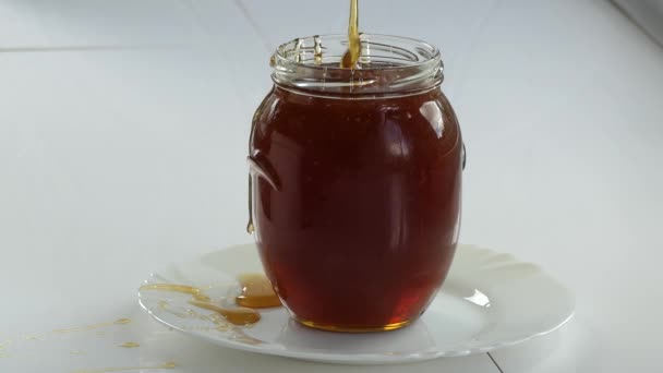 ビーガン シンプルな食べ物 白いテーブルの上に立つ透明なガラス瓶の中に薄いストリームに蜂蜜が流れます 閉じろ — ストック動画