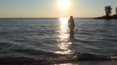 Gün batımında sahilde bir kız. Elbise giymiş güzel vücutlu bir kız gün batımında güneşli yoldan geçerek sudan çıkar. Ob Nehri, Sibirya. Yavaş çekim.