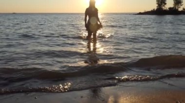 Gün batımında sahilde bir kız. Odaklanamıyorum. Elbise giymiş güzel vücutlu bir kız gün batımında güneşli yoldan geçerek sudan çıkar. Ob Nehri, Sibirya. Yavaş çekim.