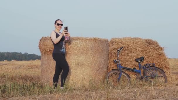 自行车 一位漂亮的骑自行车运动的女孩在收获的麦田里的稻草堆旁边看她的手机 西伯利亚 — 图库视频影像