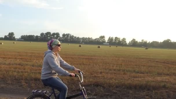 自転車だ スカーフを被った自転車愛好家の少女が畑の中にねじれたわらの玉でいっぱいの田舎道を自転車で走っています — ストック動画