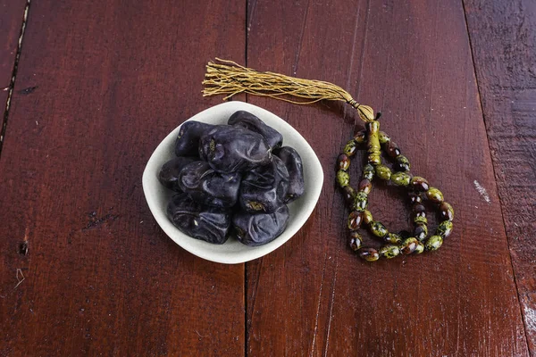 Prayer beads and Dates fruits in white bowl plate on brown wooden table surface with planks