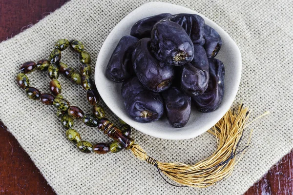 Prayer beads and Dates fruits in white bowl plate on brown wooden table with napkin
