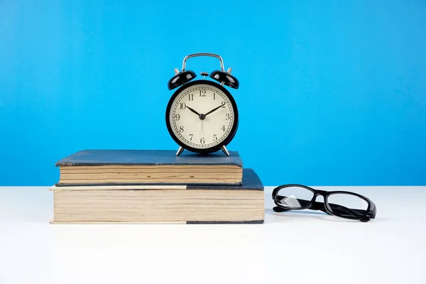 eyeglasses on closed books against blue wall and alarm vintage analog clock