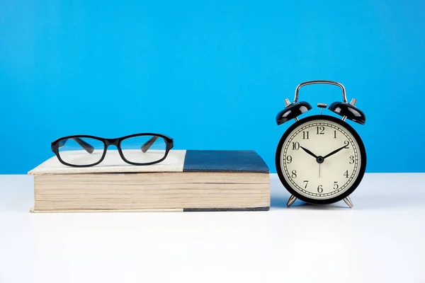 black frame eyeglasses on closed book against blue wall and alarm vintage analog clock
