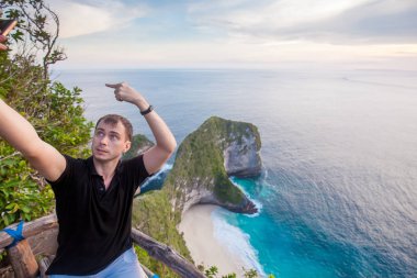 Dinozor kafası güzel tropikal plaj ve derin mavi deniz Nusa Penida Adası, günbatımı geliyor ise Endonezya'şeklinde inanılmaz yeşil taşlar ile Smartphone komik selfie yapan erkek