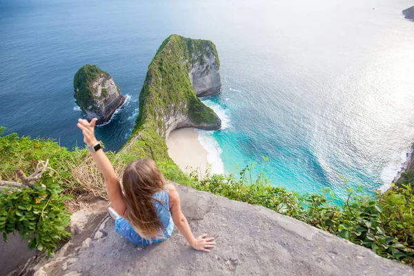 端の近くに座って 美しい熱帯のビーチとヌサ ペニダ島 インドネシアの深い青色の海の恐竜の頭のような形をした信じられないほどの緑色岩を探して長い髪の女 — ストック写真
