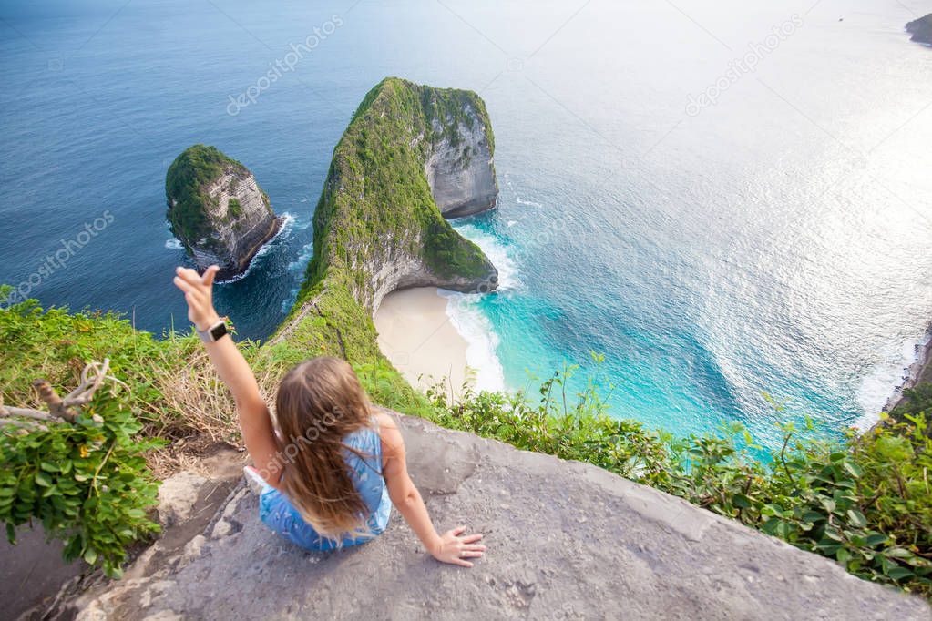 Woman with long hair sitting close to the edge and looking to the incredible green rocks shaped like dinosaur head with beautiful tropical beach and deep blue sea on Nusa Penida island, Indonesia