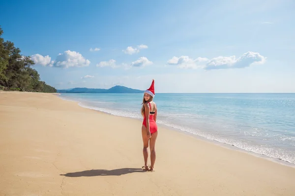 Młoda kobieta w czerwony strój kąpielowy i santa claus kapelusz stojący na plaży — Zdjęcie stockowe